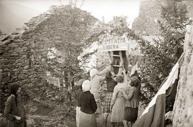 Quart (Les Trois-Villes, Avisod), 23 août 1945. 1er anniversaire des représailles nazies-fascistes du 23 août 1944 Femmes en train d’afficher un panneau avec l’inscription « W l’Italia libera » dans les décombres. © Région autonome Vallée d'Aoste - Photo Octave Bérard (fonds Bérard)